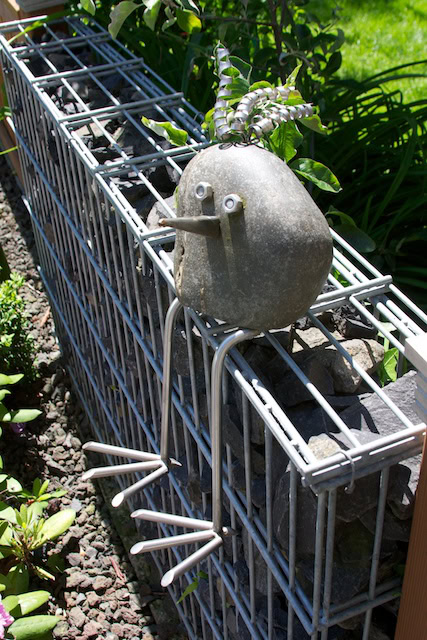Steinskulptur mit Augen und Drahtbeinen im Garten